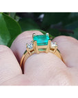 Gold ring with a natural emerald on a finger against a blurred green background.