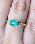 Yellow gold emerald ring on a person's finger against a blurred natural background.