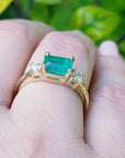 A yellow gold emerald ring three stone on a hand against a blurred green background