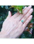 Hand wearing an emerald three stone ring against a green leafy background.
