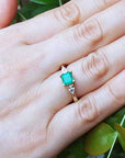 Hand wearing a ring with an emerald stone against a green leaf background.