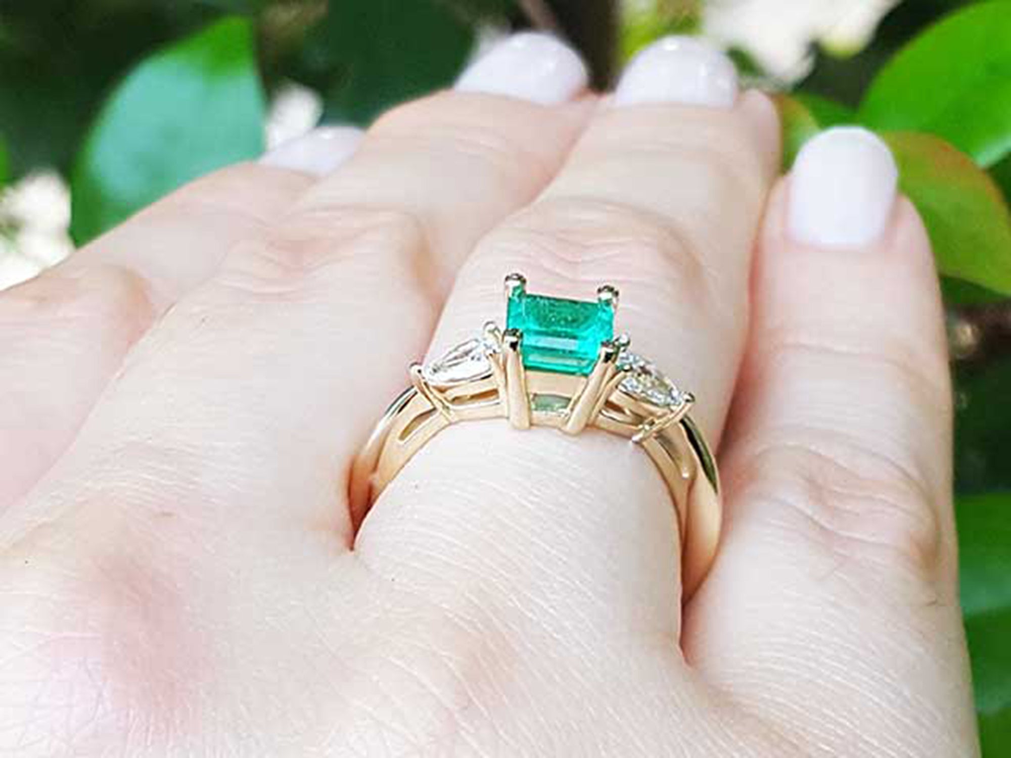 Gold ring with a natural emerald on a hand against a blurred green leafy background.