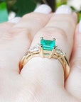 Gold ring with a natural emerald on a hand against a blurred green leafy background.