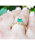 Gold ring with a natural emerald on a hand against a blurred green leafy background.