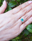 Hand wearing a lady's emerald three stone ring round cut on a green leafy background.