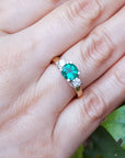 Ring with a round emerald and side diamonds on a hand against a leafy background.