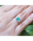 Ring with a round emerald and side diamonds on a hand against a leafy background.