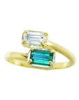 A yellow gold ring featuring two emerald-cut gemstones, with a larger green emerald and a smaller clear baguette stone above it, set against a plain white background.