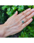 A 18k white gold toi et moi ring with a half-moon natural colombian emerald worn on a person's ring finger, shown against a background of green outdoor foliage.