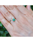 A close-up of a hand wearing a You and Me 18k gold ring with a natural emerald and diamond, set against a blurred green outdoor background.