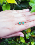 Hand displaying a pair of natural tulip emerald stud earrings against a green leafy background.