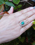 Hand wearing a vintage emerald and diamond ring against a green leafy background.