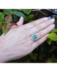 Hand wearing a vintage emerald and diamond ring against a green leafy background.