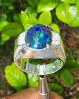 Silver ring for men with a blue-black opal held against a green leafy background