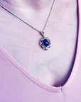 Silver pendant with an oval, deep blue gemstone, surrounded by intricate silver scrollwork, worn on a delicate chain.