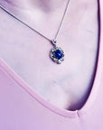 Silver pendant with an oval, deep blue gemstone, surrounded by intricate silver scrollwork, worn on a delicate chain.