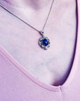 Silver pendant with an oval, deep blue gemstone, surrounded by intricate silver scrollwork, worn on a delicate chain.