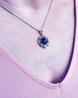 Silver pendant with an oval, deep blue gemstone, surrounded by intricate silver scrollwork, worn on a delicate chain.