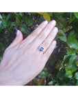 Hand wearing a white gold ring with a blue natural sapphire against a green leafy background