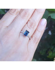Hand wearing a white gold ring with a blue sapphire gemstone against a blurred green leafy background
