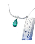 White gold necklace with a pear-shaped emerald and diamond-studded wavy bar, next to a ruler.
