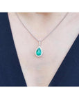 Pear-shaped deep green emerald pendant with a diamond halo on an 18K white gold chain, worn on a person.