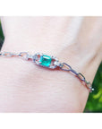 A white gold paperclip bracelet with a square emerald on a wrist against a blurred natural background.