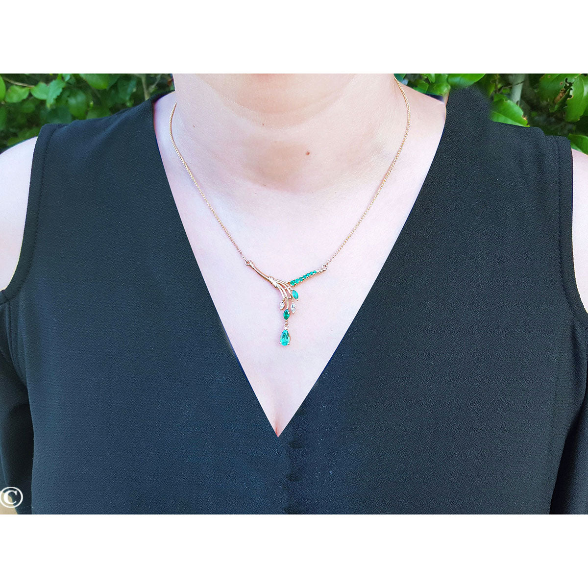 Yellow gold necklace with natural emeralds on a woman collar wearing a dark green top against a blurred green background
