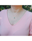Woman wearing a pink top with a necklace featuring an emerald pendant against a blurred natural background