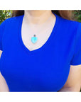 Person wearing a blue shirt with a necklace featuring a heart-shaped larimar pendant outdoors.