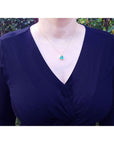 Person wearing a dark blue top with a necklace featuring a emerald heart pendant against a blurred natural background.