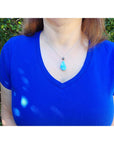Person wearing a blue shirt with a necklace featuring a larimar pendant against a green outdoor background.