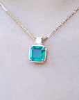 Front side of a yellow gold necklace with an Asscher cut emerald gemstone pendant on a light background