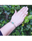Emerald bangle bracelet with natural diamonds on 14K white gold, worn on a hand against green leaves.