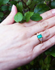 Hand wearing a yellow gold emerald ring with a leafy background.
