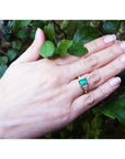 Hand wearing a yellow gold emerald ring with a leafy background.