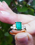 A yellow gold ring with emerald and side diamonds held between fingers.