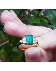 A yellow gold ring with emerald and side diamonds held between fingers.