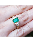 A yellow gold ring with a natural emerald and diamond accents on a finger.