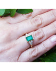 Gold ring with an emerald and diamond accents on a leafy background.