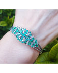A white gold emerald bangle bracelet on a wrist against a green leafy background.