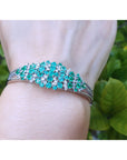 Bracelet with round cut emerald and diamonds on a wrist against a green leafy background.
