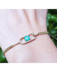 Gold bracelet with a colombian emerald on a wrist against a blurred green background.