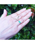14K yellow gold necklace with a baguette-cut Colombian emerald dangling from a diamond-accented V-pendant, shown worn on a person's finger with their hand resting against a background of green leaves.