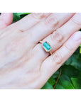 Hand wearing a ring with an emerald stone against a green leafy background.