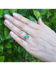 A hand wearing an emerald solitaire ring yellow gold against a green leafy background.