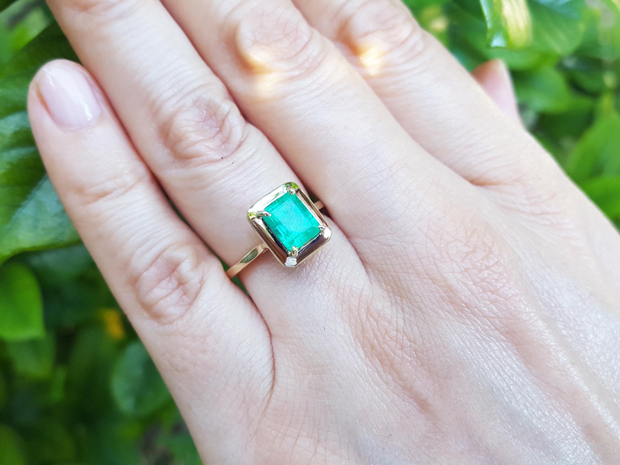 A close-up of a hand wearing a yellow gold emerald solitaire ring on a green blurry background.
