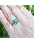 A close-up of a hand wearing a yellow gold emerald solitaire ring on a green blurry background.