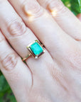 A close-up of a hand wearing a yellow gold emerald solitaire ring on a green blurry background.