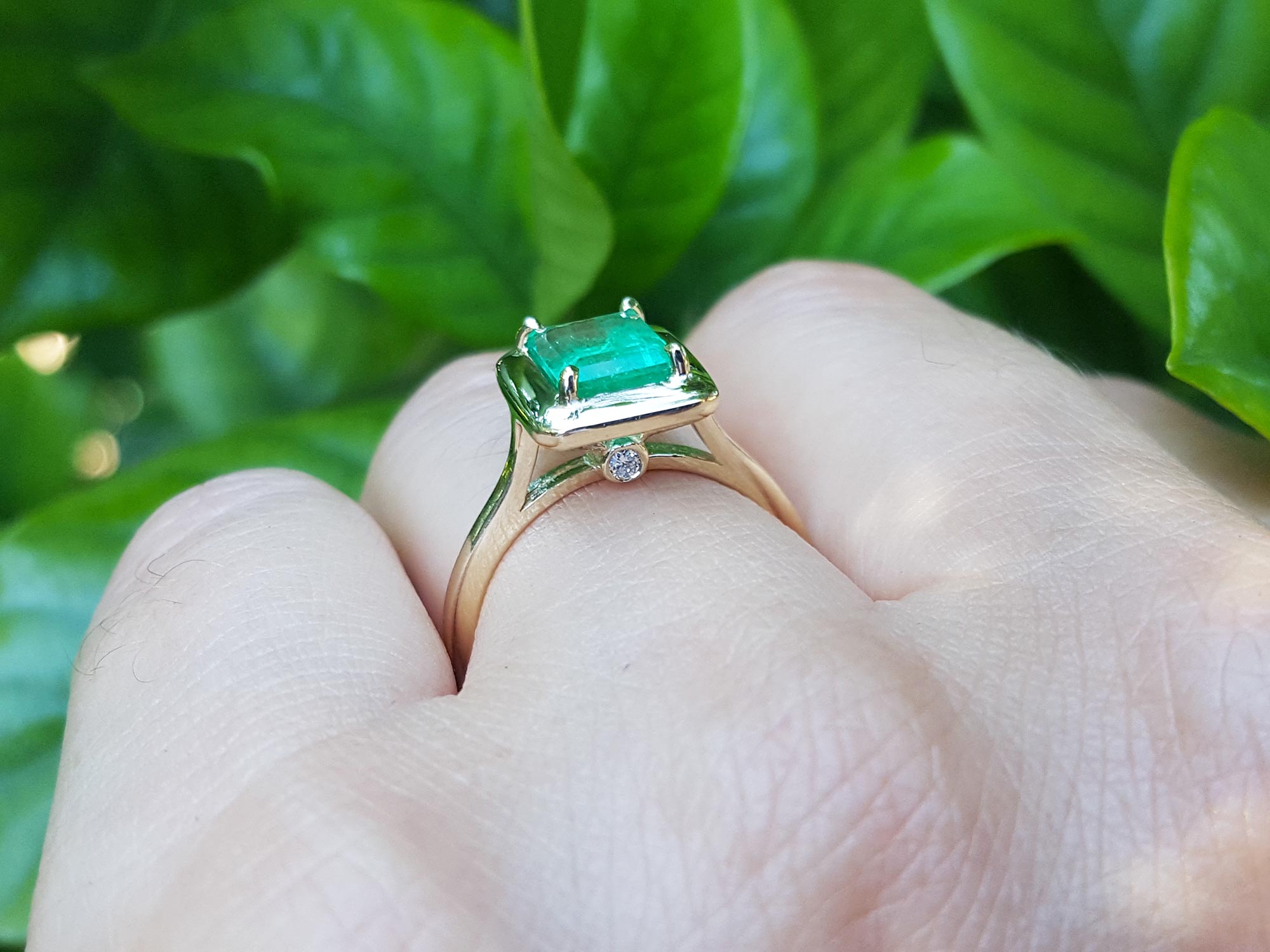 On a hand the underside view of an emerald solitaire ring in yellow gold against a leafy background.